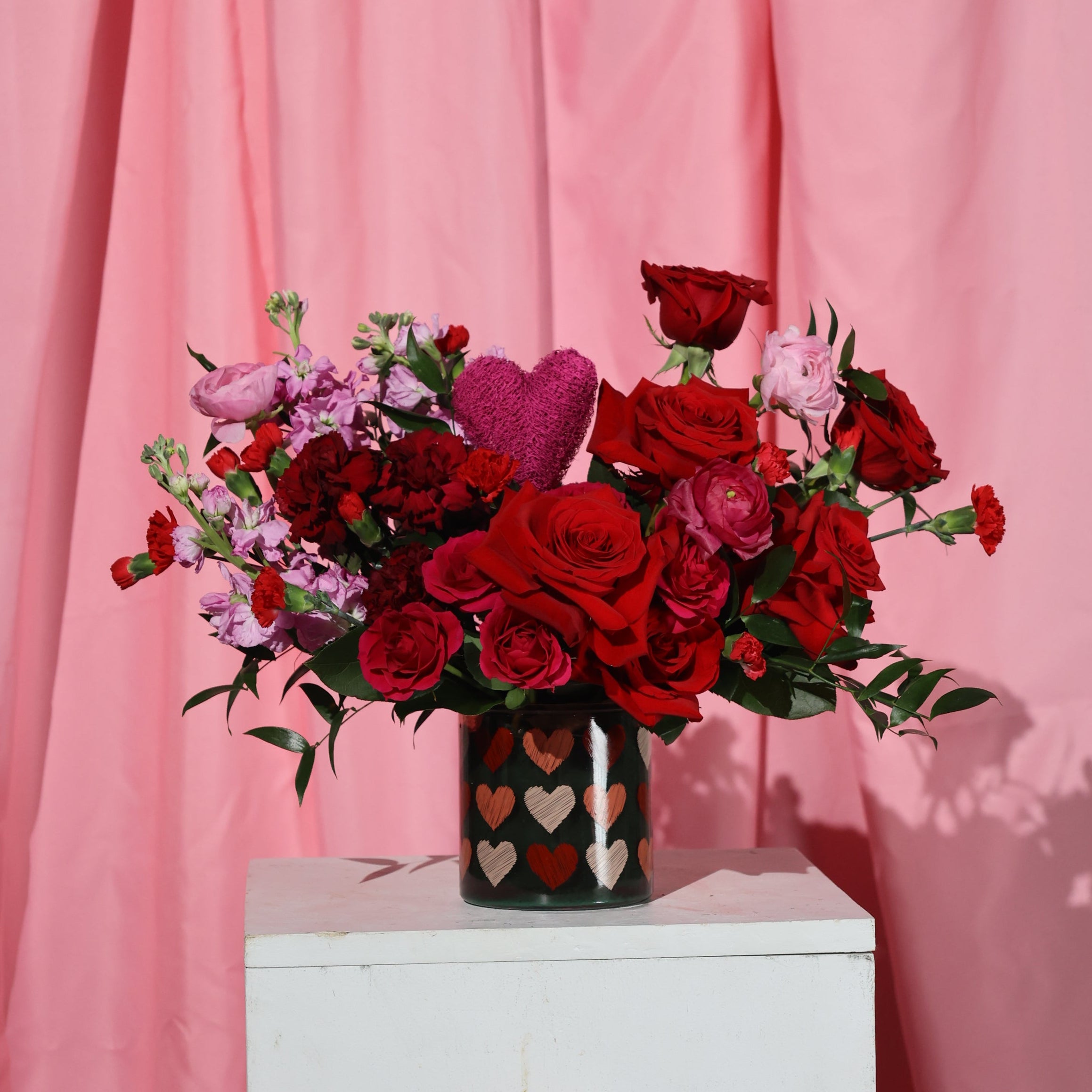 Bouquet of red and pink flowers in a vase with heart design against a pink curtain.